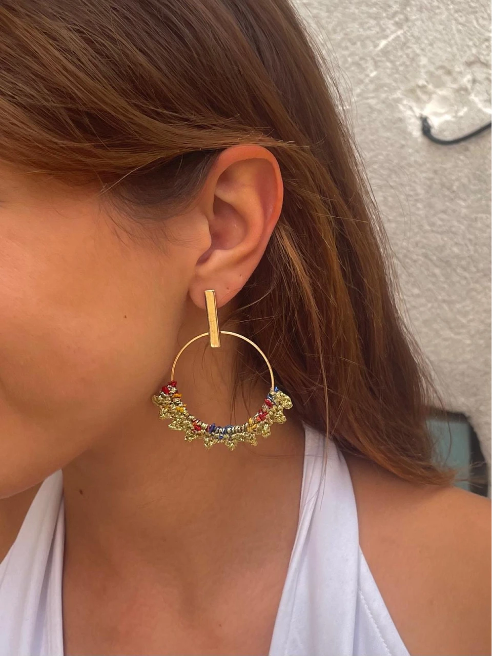 Une personne porte les Boucles d'oreilles Cora de Croch' et Chic, qui sont de grandes créoles ornées de perles colorées et d'accents dorés. Les boucles d'oreilles présentent une barre verticale distinctive reliée au créole. La personne a de longs cheveux bruns et est vêtue d'un haut blanc.