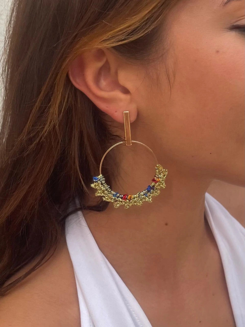 Un gros plan montre une personne portant les « Boucles d'oreilles Cora » de Croch' et Chic, composées de grandes créoles en or ornées de petites perles bleues, rouges et dorées. La boucle d'oreille est suspendue à une attache rectangulaire en or. La personne a les cheveux châtain clair et est vêtue d'un haut blanc.